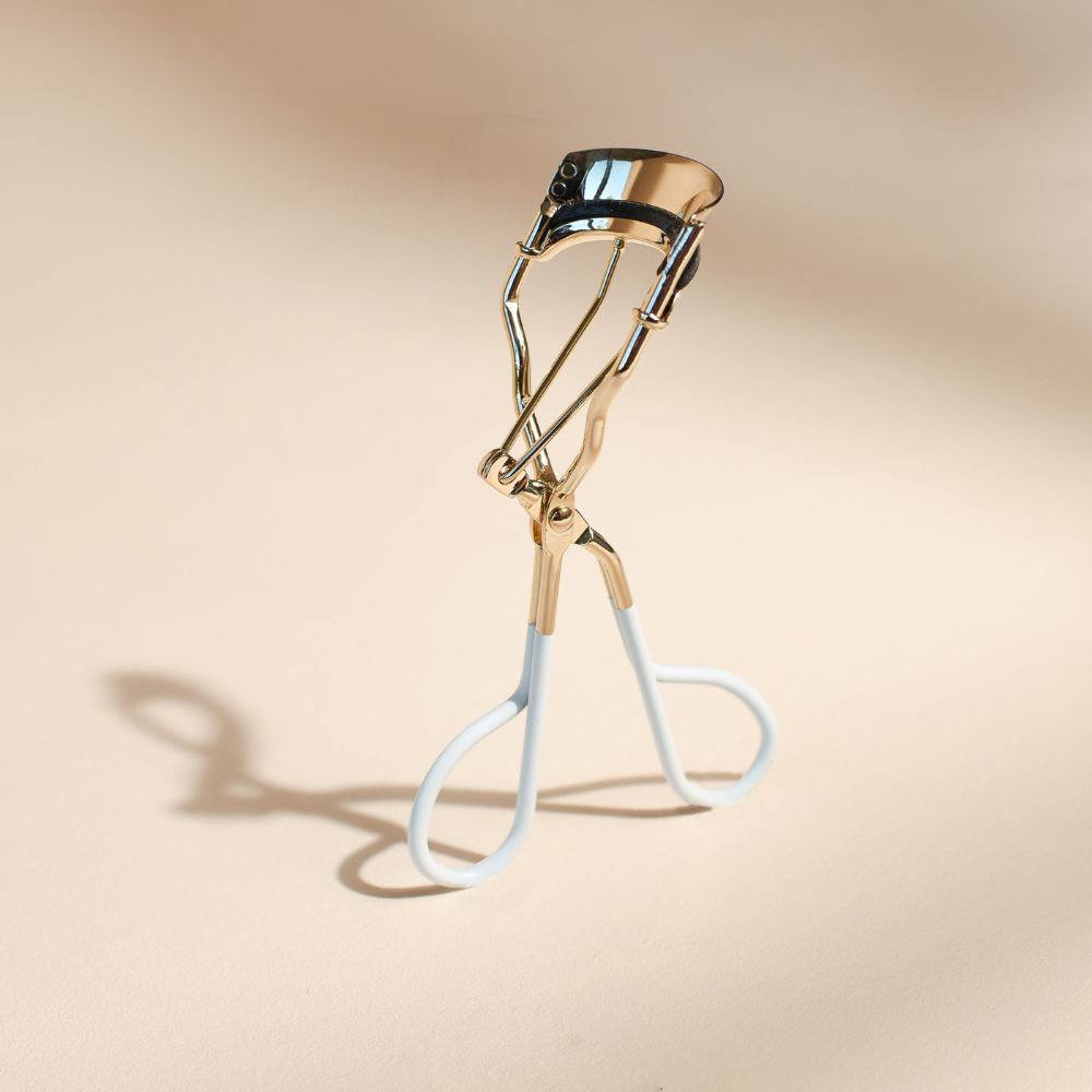 Recourbe-cils Bachca en métal doré avec poignées blanches, posé debout sur un fond beige avec une ombre élégante.