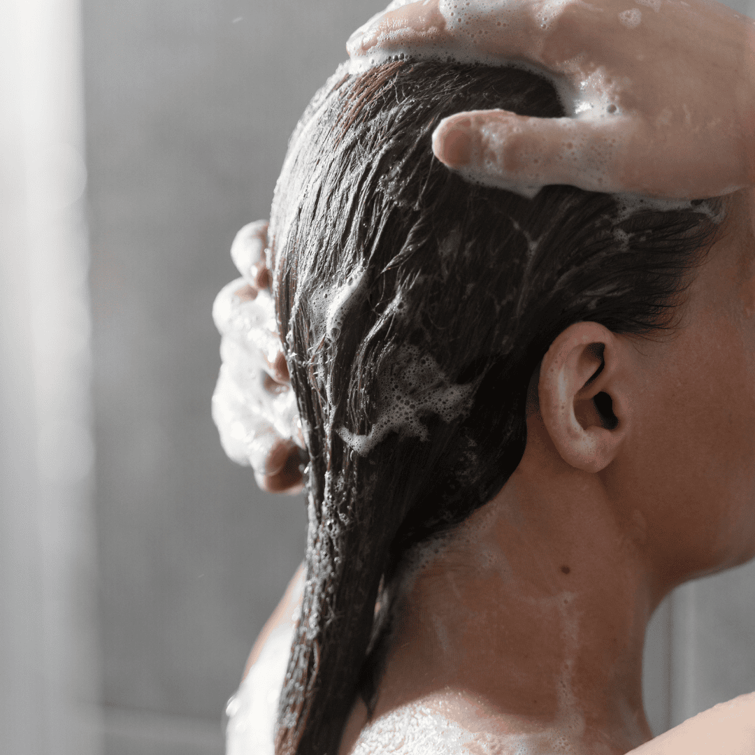 Femme sous la douche lavant ses cheveux mouillés avec un shampooing moussant, moment de soin capillaire et détente à domicile.