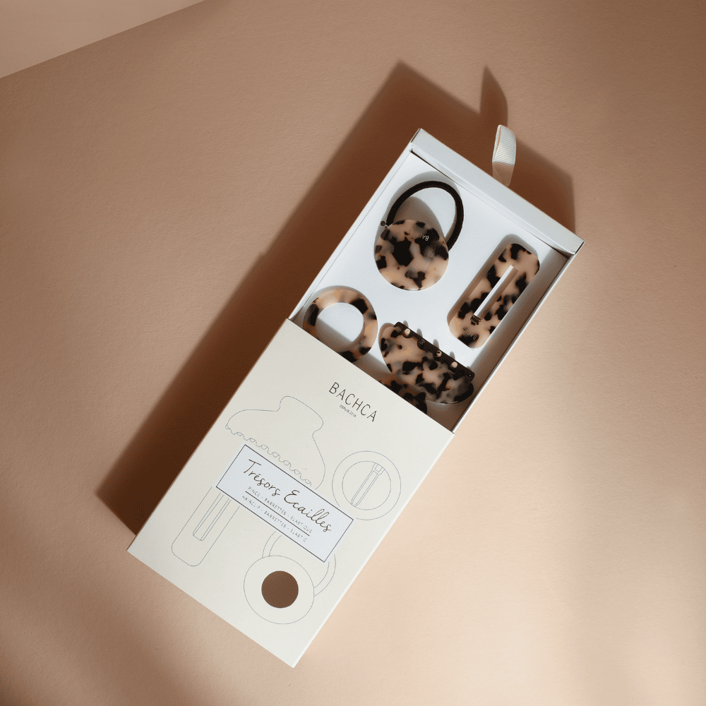 Coffret d’accessoires cheveux en écaille de la marque Bachca : pince, barrette, barrette ronde et élastique plaque, présentés sur fond rose pâle.