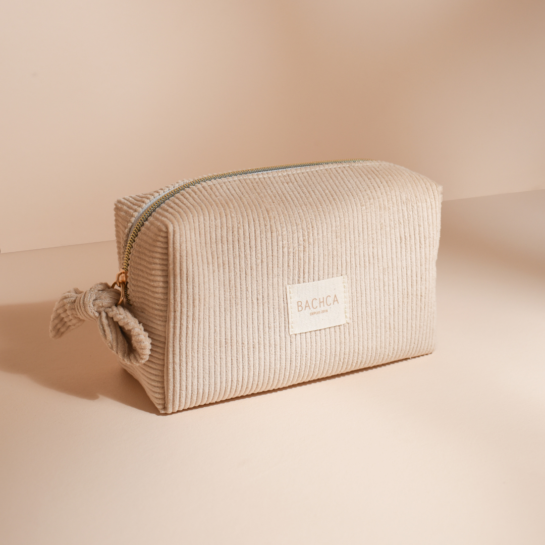 Trousse de maquillage en velours écru BACHCA avec zip doré, posée sur fond beige naturel.