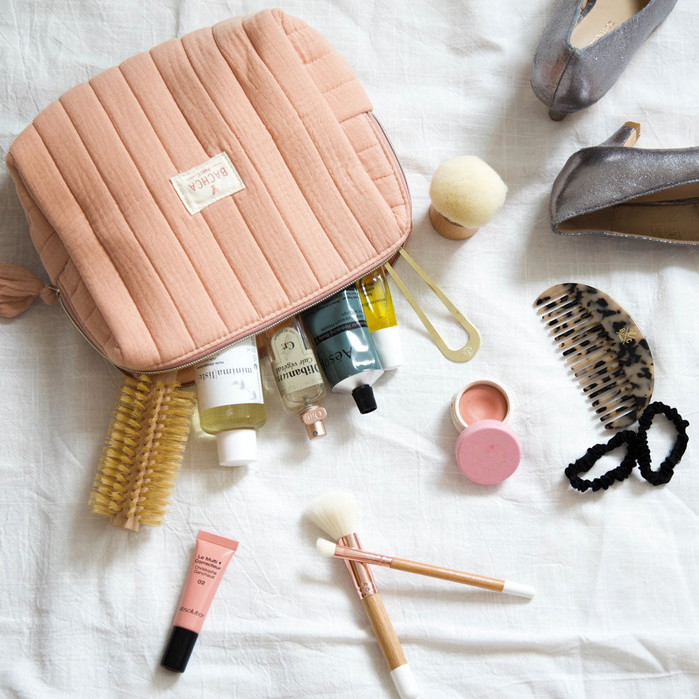 Trousse de toilette Bachca rose poudré ouverte avec brosse ronde, peigne écaille, pinceaux maquillage et soins beauté disposés sur un drap blanc.