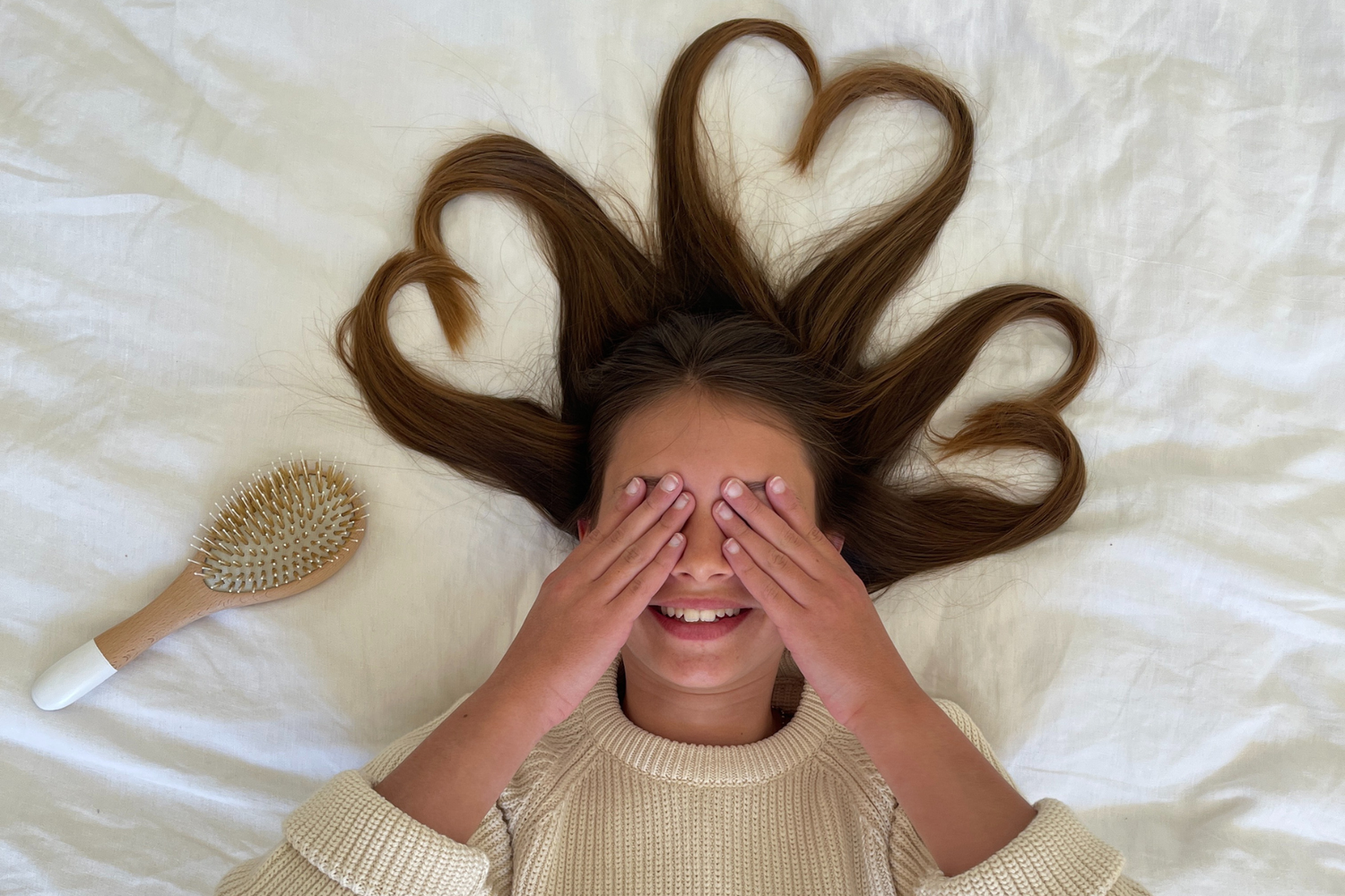 Petite fille se cachant les yeux avec ses cheveux formant des coeurs sur un fond blanc, une brosse à cheveux de la marque Bachca se place à côté d'elle.