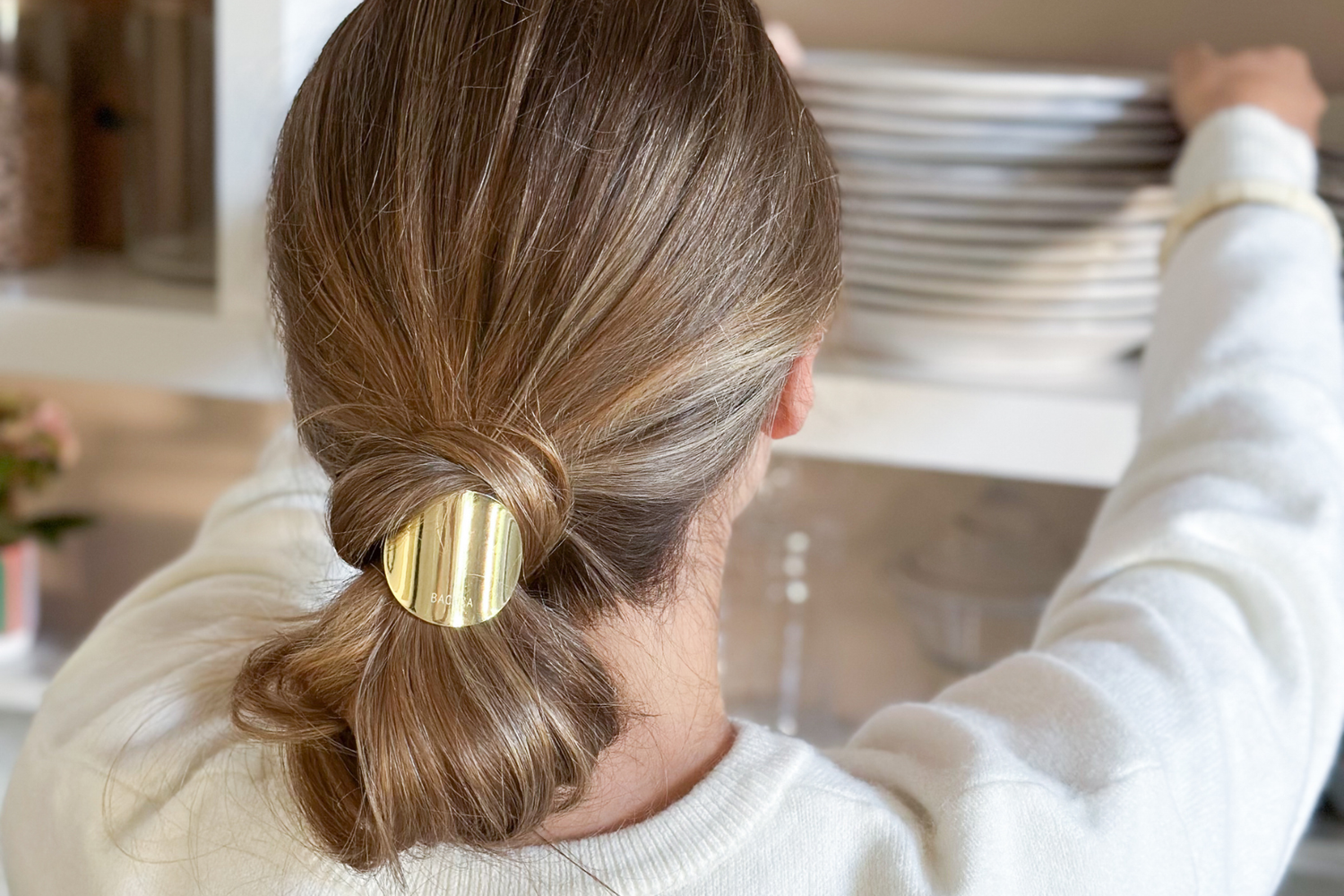 Jeune femme de dos avec des cheveux blonds attachés en chignon bas à l'aide d'un élastique plaque doré de la marque Bachca.