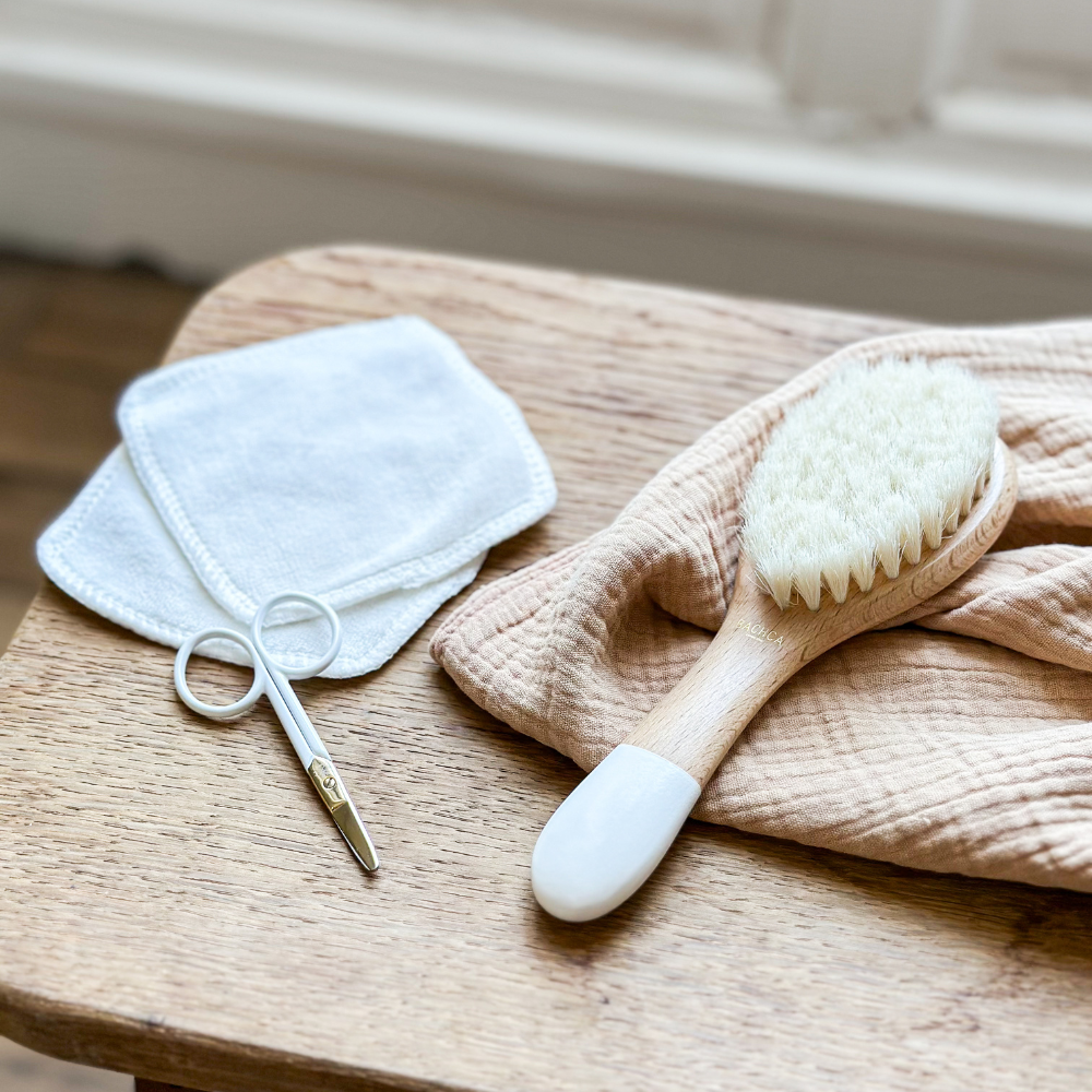 Brosse à cheveux pour nourrisson en soie de chèvre posée sur un linge beige, un ciseaux bébé et deux cotons réutilisables sont posés à côté.