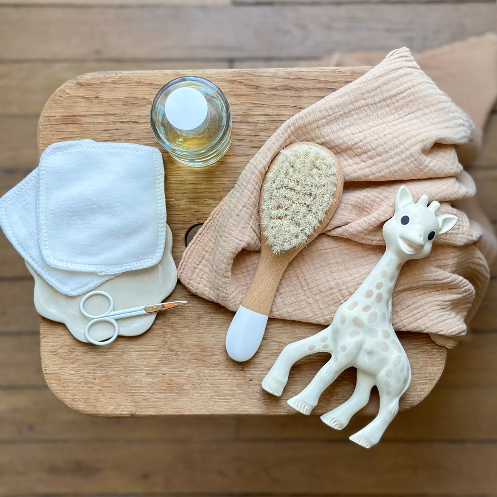 Brosse à cheveux pour nourrisson en soie de chèvre posée sur un tabouret en bois accompagné d'un lot de deux cotons et d'un ciseaux à ongles bébé.