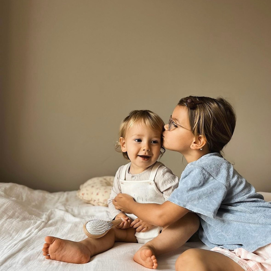 Deux enfants assis sur un lit, la plus grande embrassant le plus jeune qui tient une brosse à cheveux Bachca.