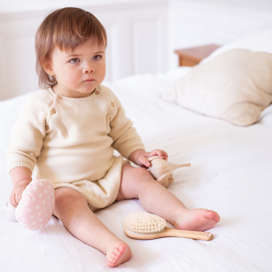 Bébé assis sur un lit blanc, tenant des jouets, avec une brosse bébé en soie de chèvre Bachca posée à ses pieds.