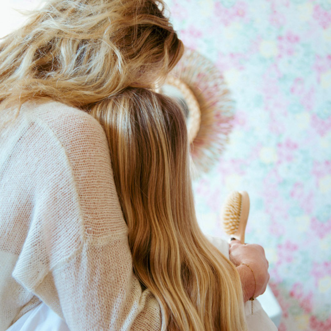 Mère et fille aux cheveux blonds enlacées, la fille tenant une brosse à cheveux Bachca en bois.