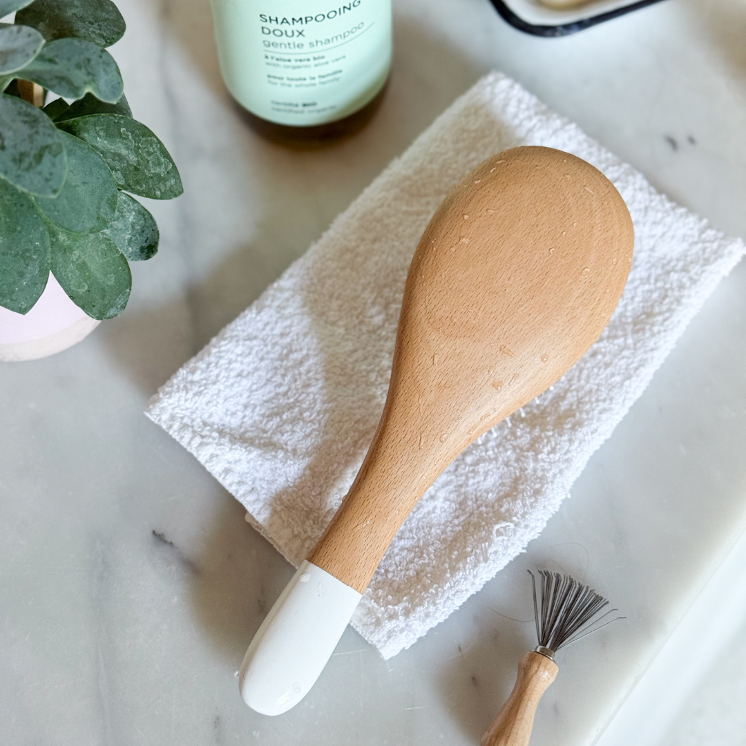 Brosse à cheveux posée sur une serviette blanche avec un peigne nettoyant pour brosse, sur un fond épurée de salle de bain.