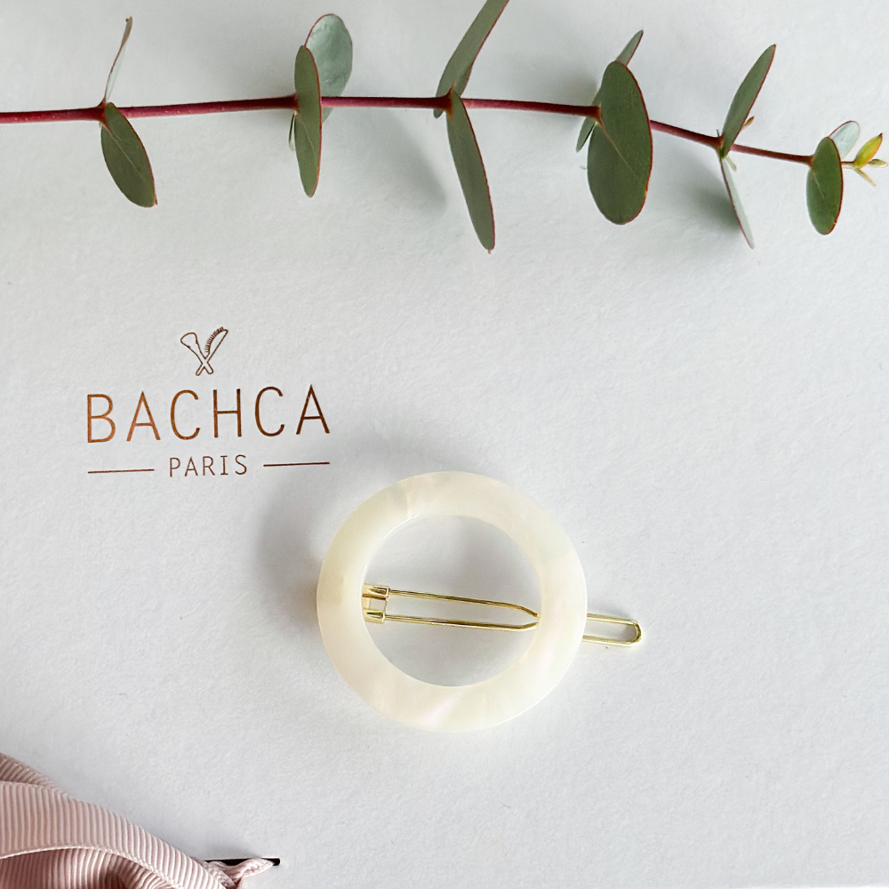 Barrette ronde nacrée Bachca posée sur fond clair avec logo de la marque et branche d’eucalyptus, accessoire chic et naturel pour cheveux.
