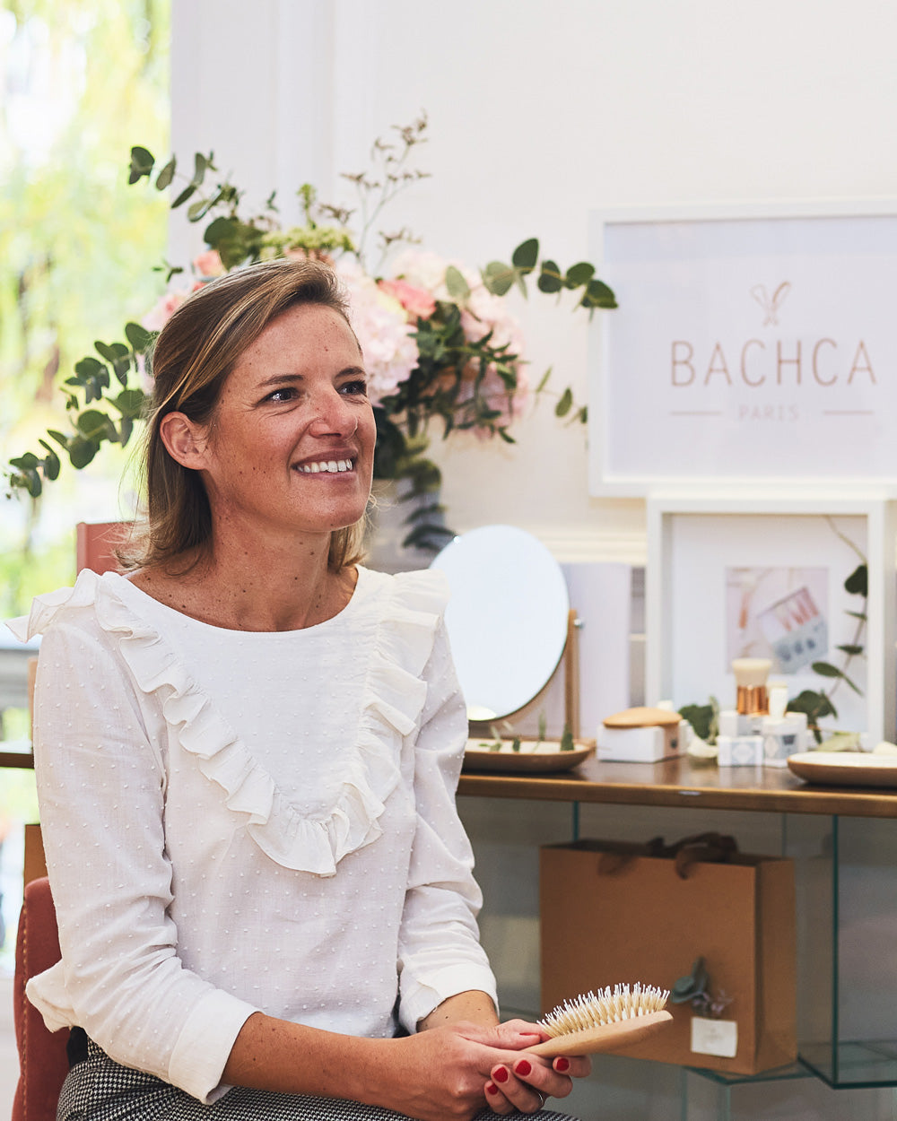 Femme souriante tenant une brosse à cheveux Bachca, assise dans un décor raffiné avec des fleurs et des produits de la marque exposés en arrière-plan.