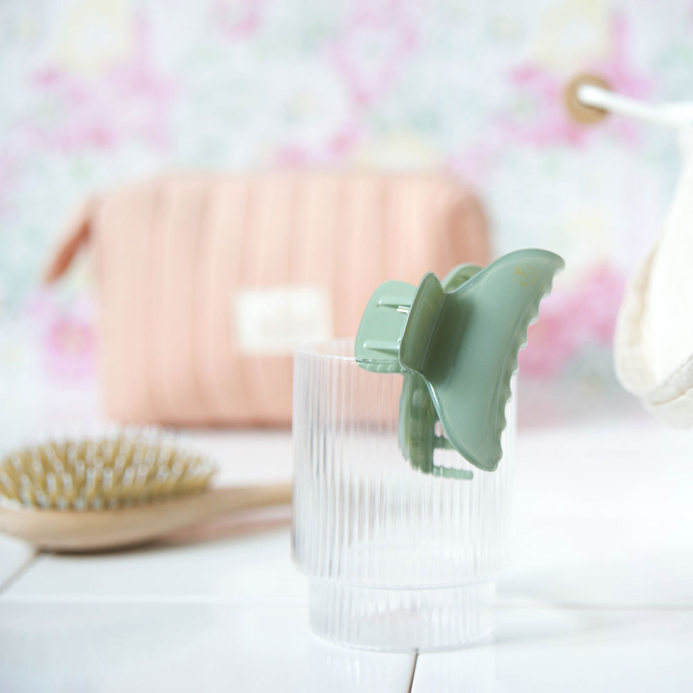 Pince à cheveux croco Bachca vert eucalyptus posée sur un verre, avec trousse et brosse en arrière-plan.