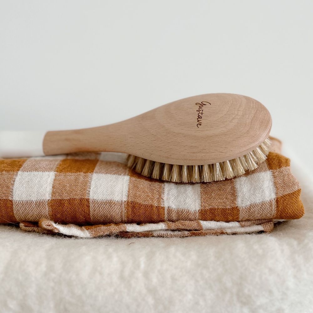 Brosse bébé en bois de hêtre Bachca posée sur un lange à carreaux orangés et blancs, ambiance douce et minimaliste.