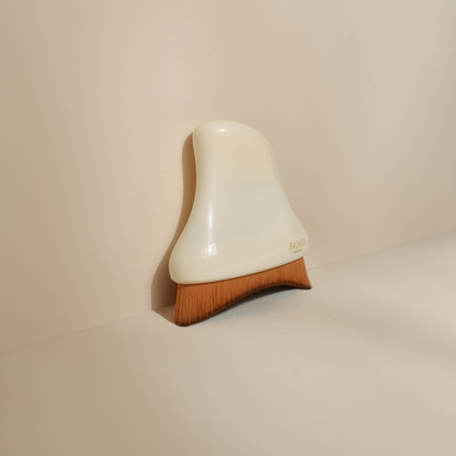 Photo studio sur fond beige, la brosse visage de drainage lymphatique Bachca est posée sur le fond, on peut ainsi voir sa forme rectangulaire et ses poils synthétiques.