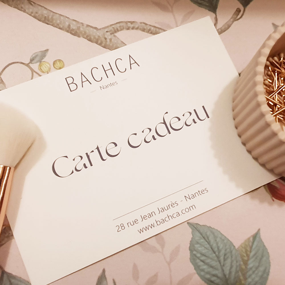 Une carte cadeau Bachca posée sur un fond fleuri, accompagnée d’un pinceau et d’un pot de pinces dorées, prête à offrir.