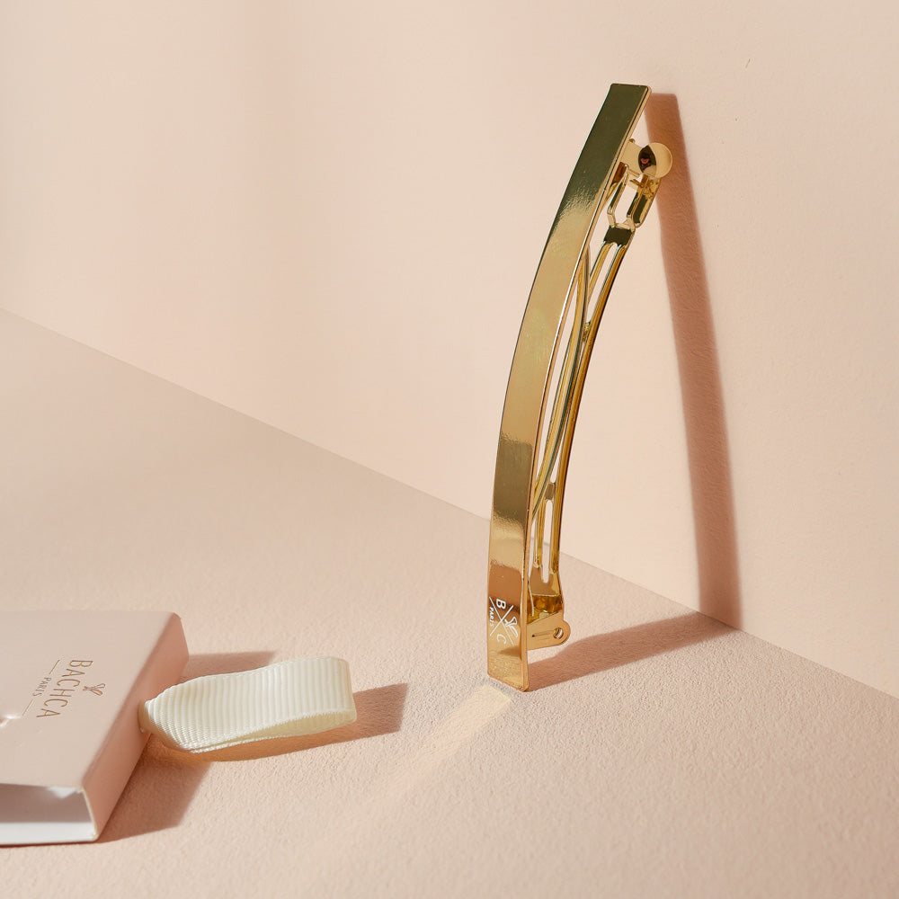 Barrette longue Bachca en métal doré, présentée debout avec son ombre portée, à côté de son support cartonné rose poudré, photographiée sur une surface beige.