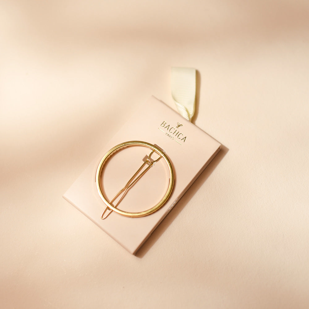Barrette ronde dorée Bachca présentée sur son carton beige rosé, posée sur un fond uni dans les mêmes tons poudrés.
