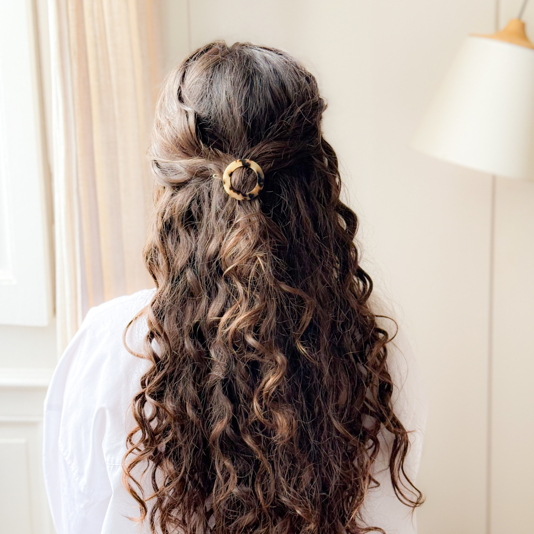 Cheveux longs et bouclés attachés avec une barrette ronde motif écaille Bachca, coiffure semi-attachée dans un intérieur lumineux.