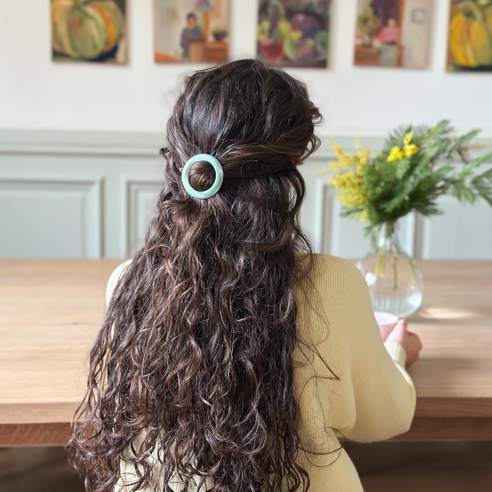 Femme de dos aux cheveux longs bouclés attachés en demi-queue à l'aide d'une barrette ronde vert d'eau de la marque Bachca. 