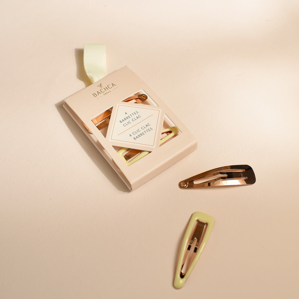 Boîte de quatre barrettes clic-clac Bachca posée sur un fond beige, avec deux barrettes dorées et roses disposées à côté de l’étui.