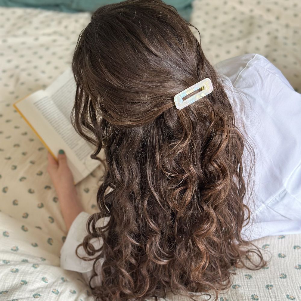 Femme allongée lisant un livre, cheveux longs et bouclés attachés avec une barrette rectangulaire nacrée Bachca, finition irisée et élégante.