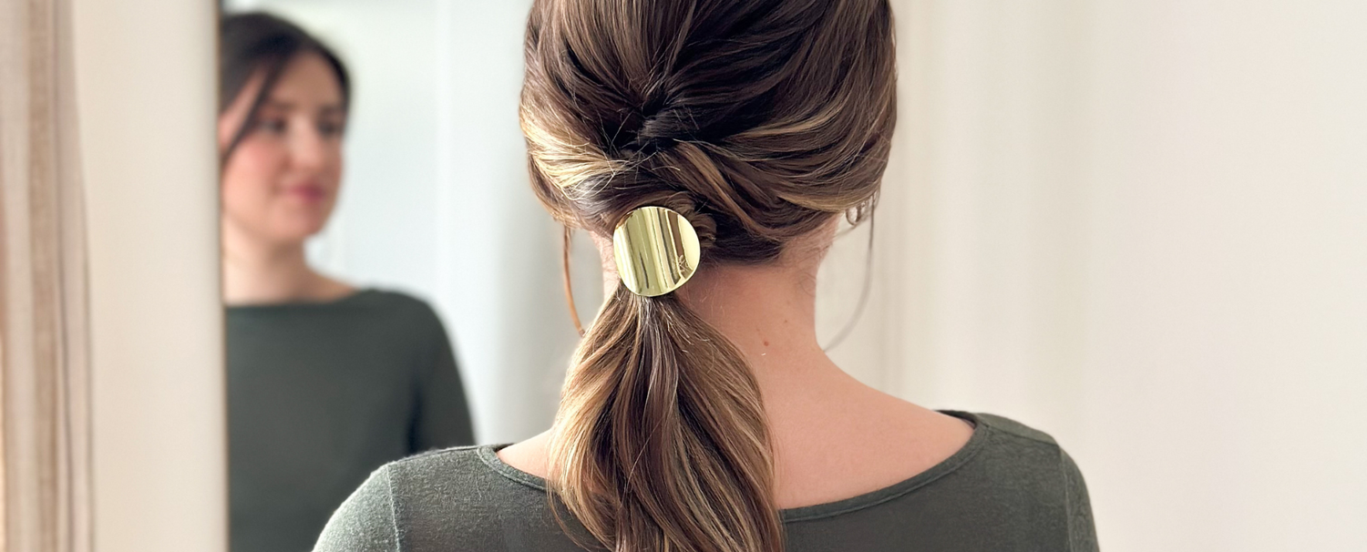 Femme de dos portant une queue de cheval basse travaillé attachés à l'aide de l'élastique plaque doré de la marque Bachca. 