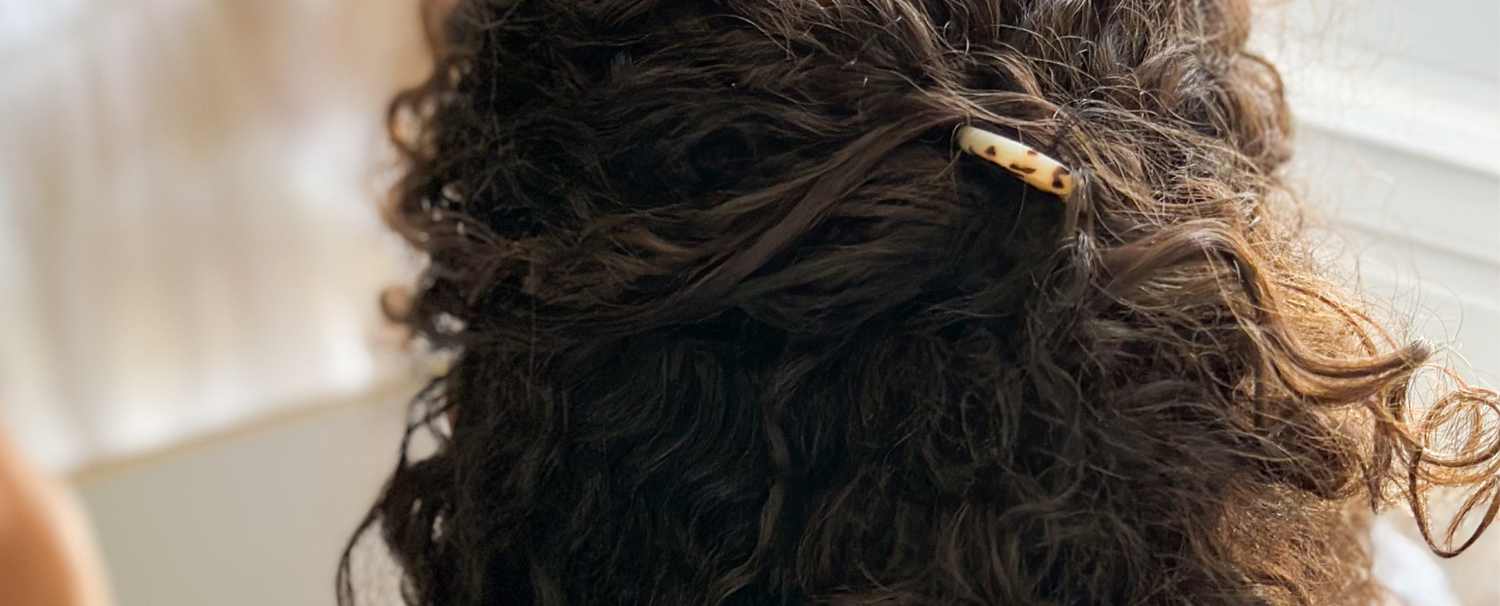 Gros plan sur des cheveux bruns bouclés attachés en demi-queue par une épingle à chignon écaille de la marque Bachca. 