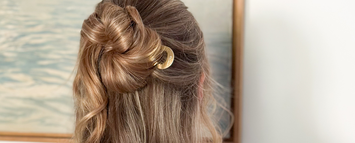 Femme aux cheveux blonds de dos portant un chignon en demi-queue attaché à l'aide d'un pic chignon doré de la marque Bachca. 