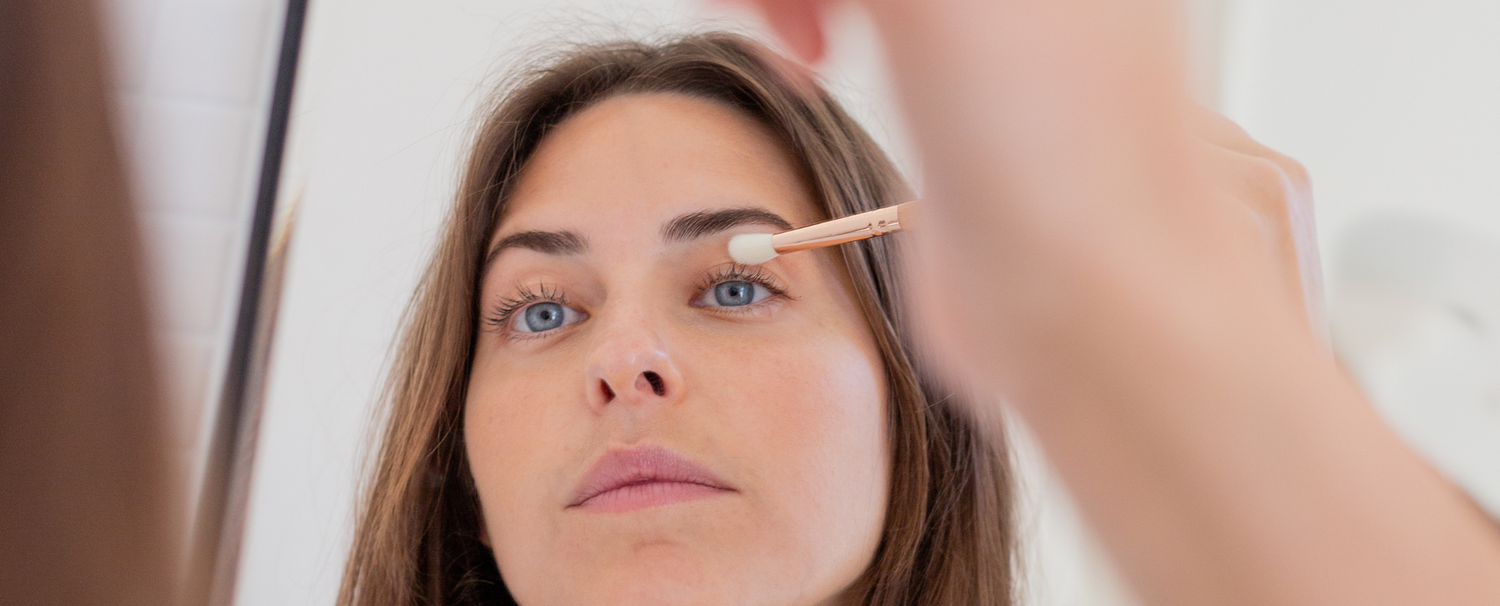 Femme aux yeux bleus dans le reflet d'un miroir utilisant un pinceau à maquillage de la marque Bachca. 