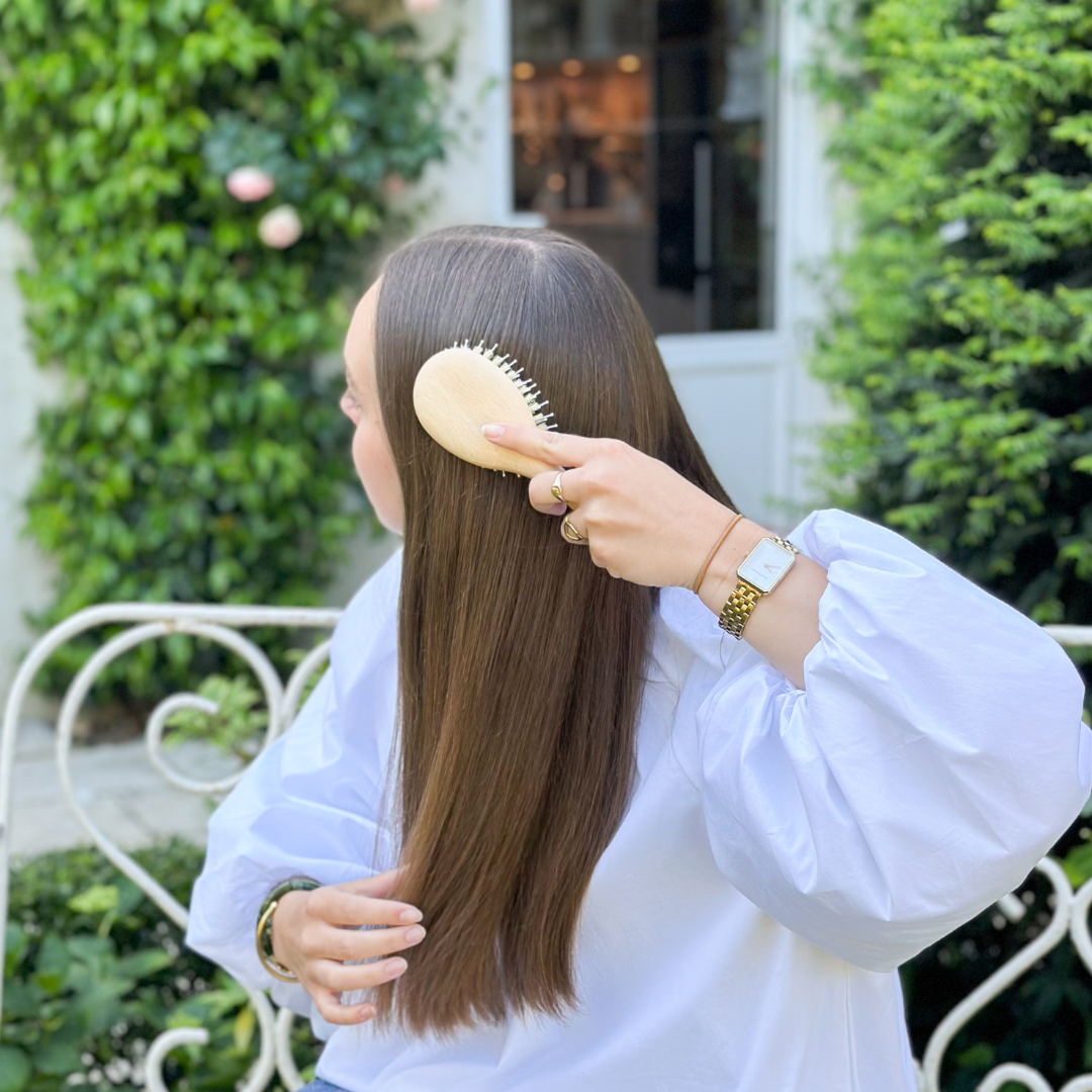 Brossage des cheveux longs et lisses avec une brosse Bachca en bois naturel, sur un banc blanc dans un jardin verdoyant.