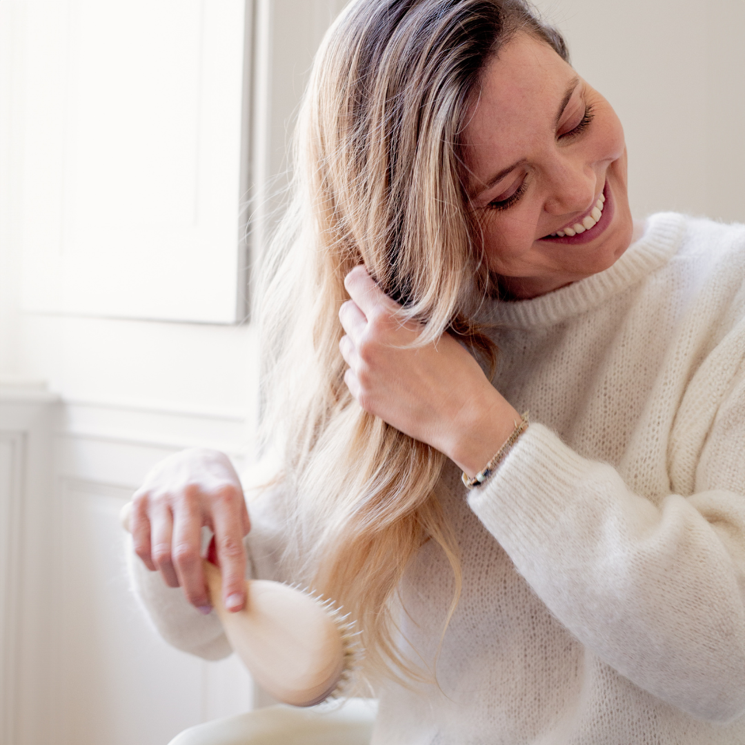 Femme souriante se brossant les cheveux blonds avec une brosse Bachca en bois, dans une pièce lumineuse au style épuré.
