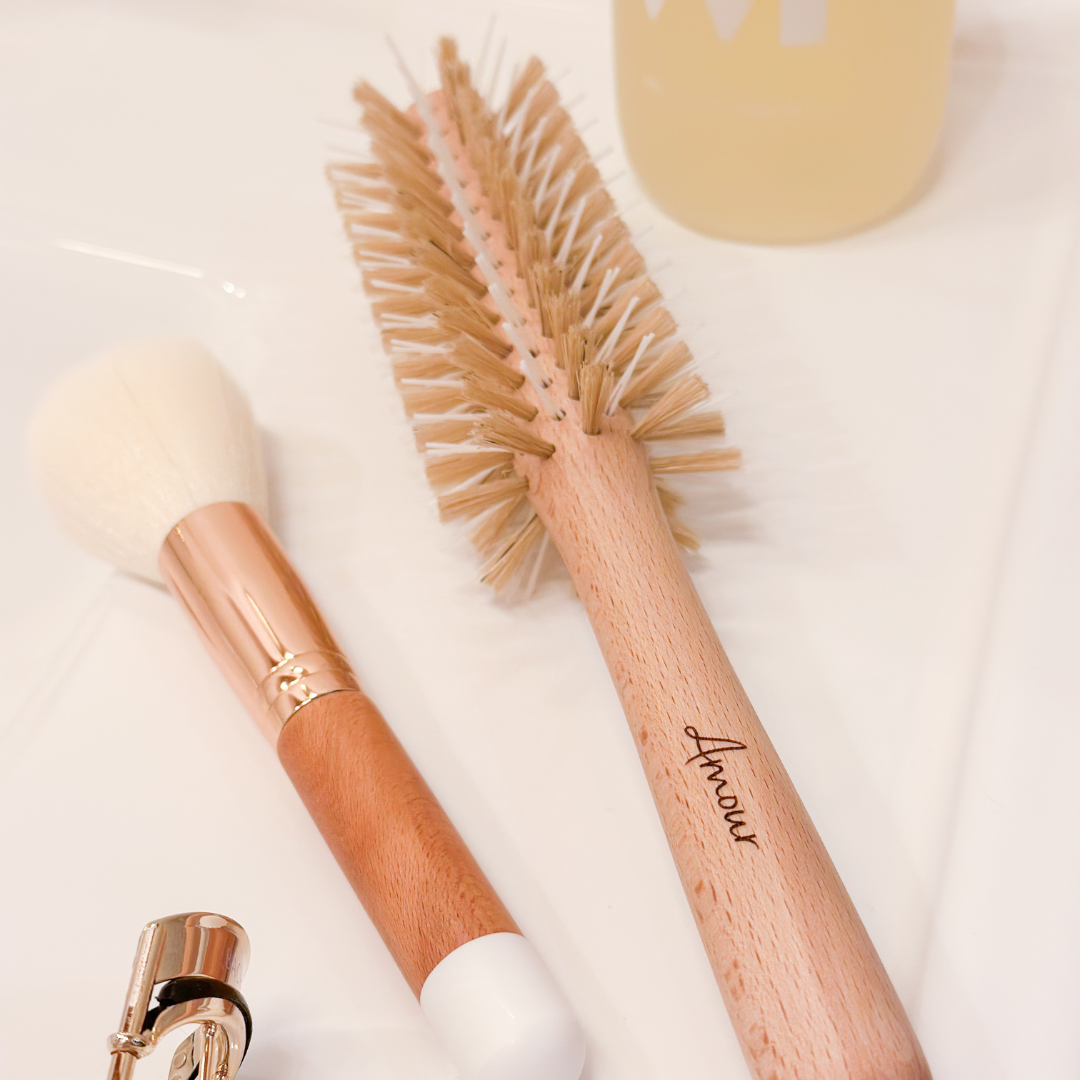 Brosse à cheveux pour brushing fabriquée en France avec picots en nylon et en sanglier de la marque Bachca. La brosse est gravée "Amour" sur son manche en bois. La brosse est posée sur un lavabo blanc avec un pinceau à maquillage posé à côté.