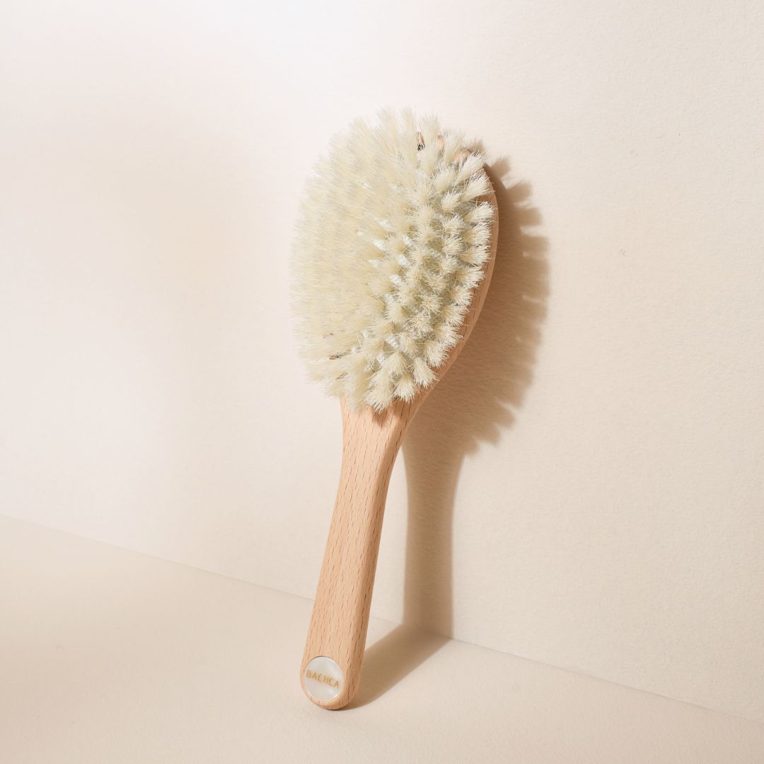 Brosse à cheveux pour bébé Bachca en bois de hêtre, à poils très souples blancs, appuyée verticalement contre un mur beige avec une ombre douce en éventail.