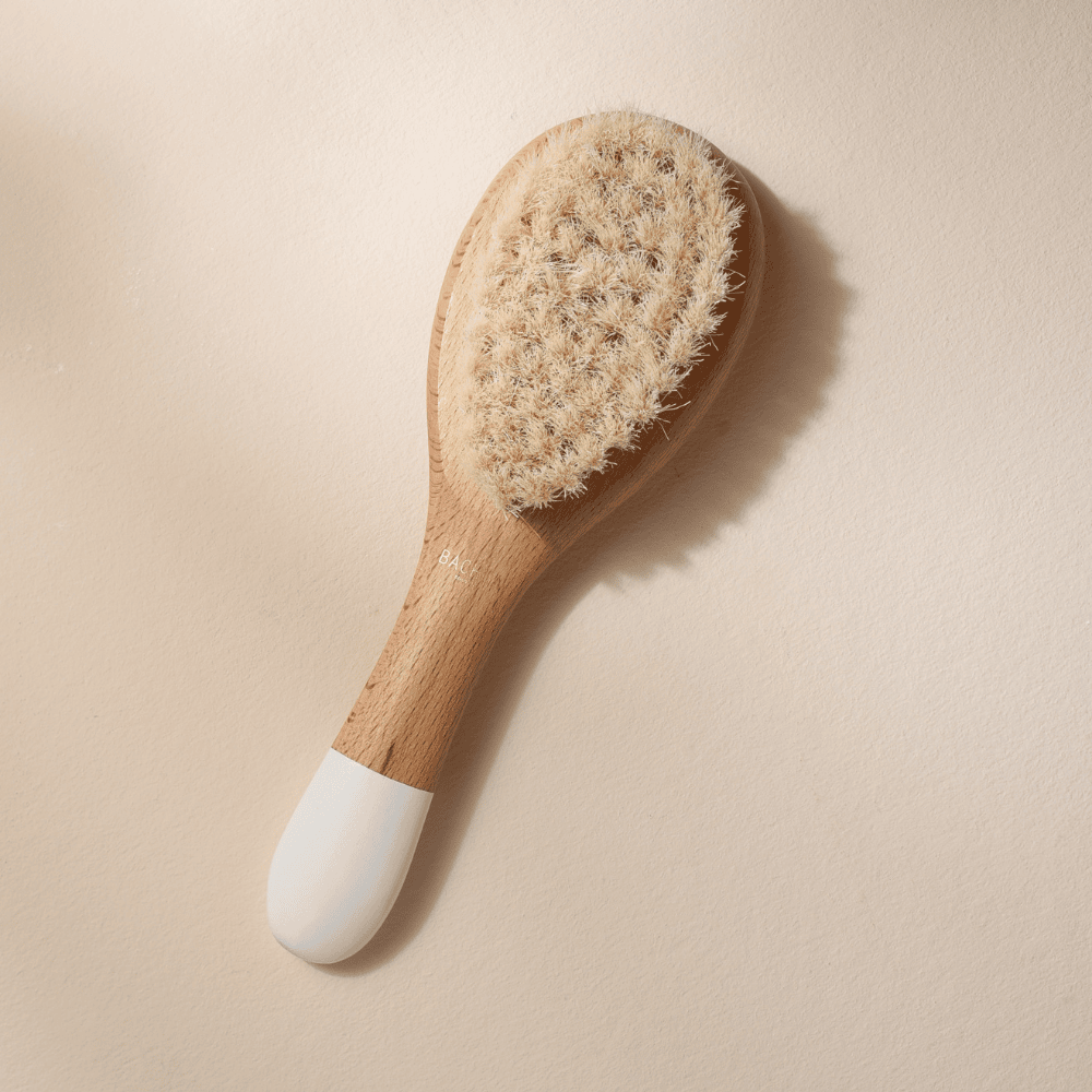 Petite brosse à cheveux de la marque Bachca. En soie de chèvre pour les cheveux de bébé et avec un manche en bois. Produit sur fond rose pâle.
