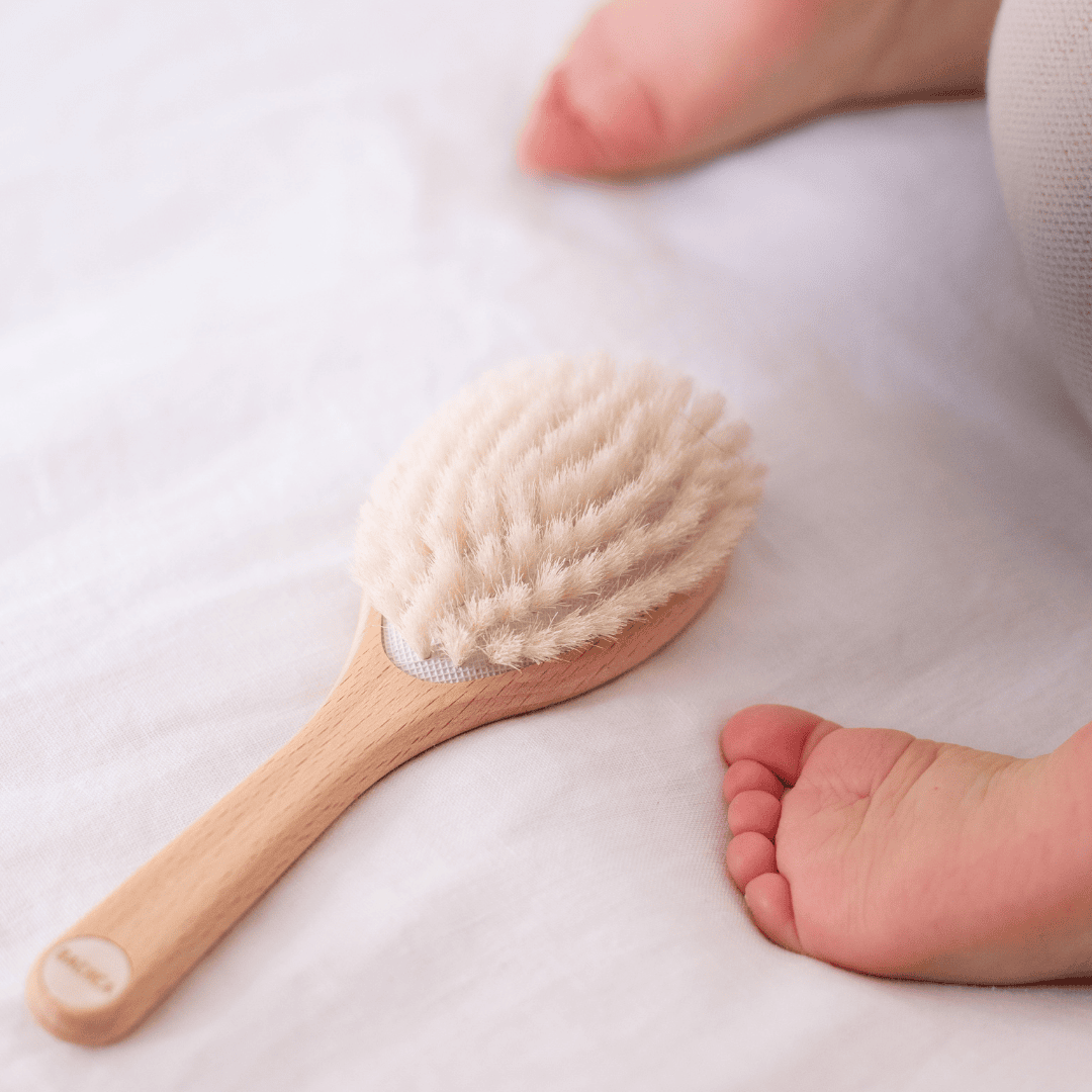 Brosse bébé fabriquée en France Bachca en bois et poils de chèvre posée sur un drap blanc, à côté des pieds d’un nourrisson allongé.