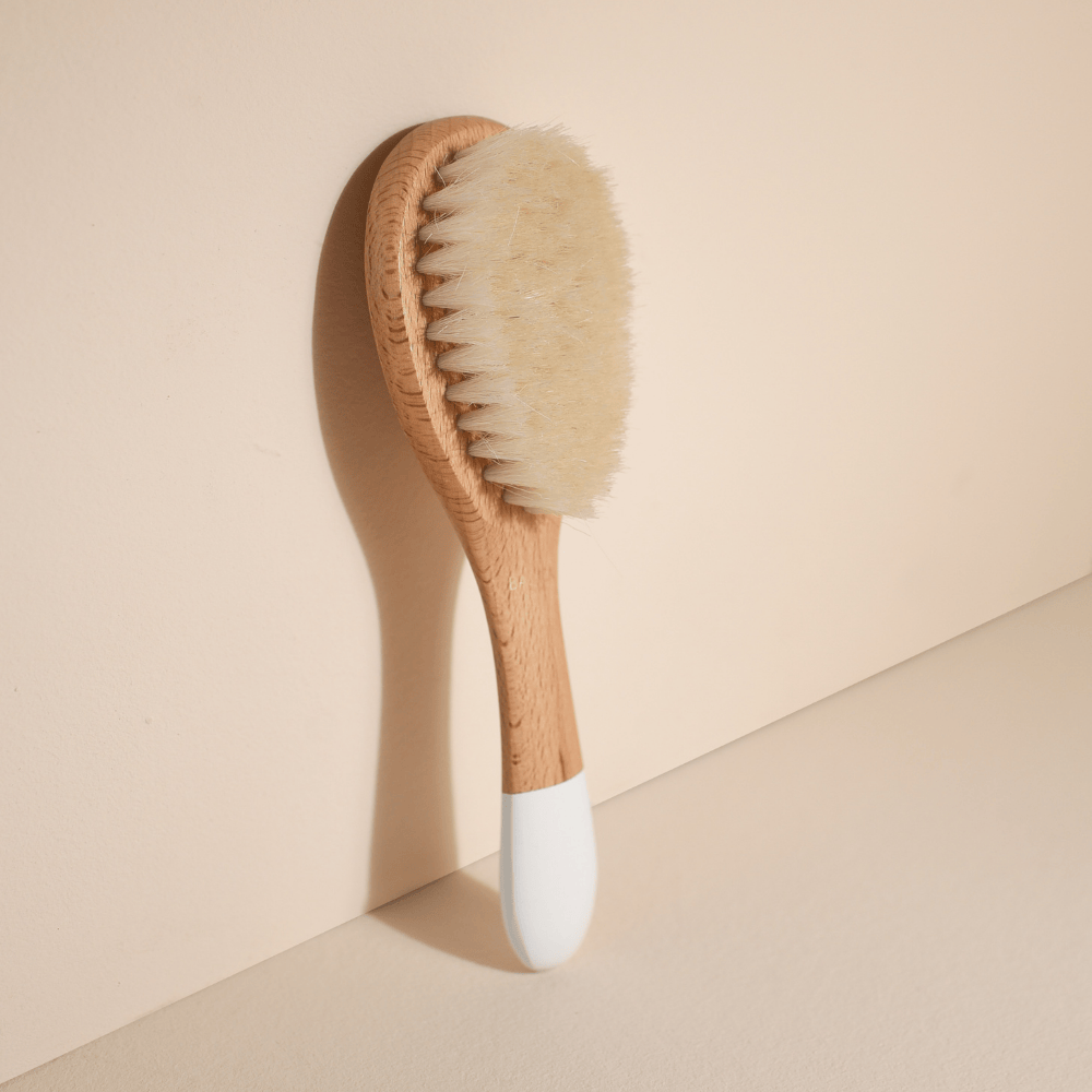 Petite brosse à cheveux de la marque Bachca. En soie de chèvre pour les cheveux de bébé et avec un manche en bois. Produit adossé sur fond rose pâle.