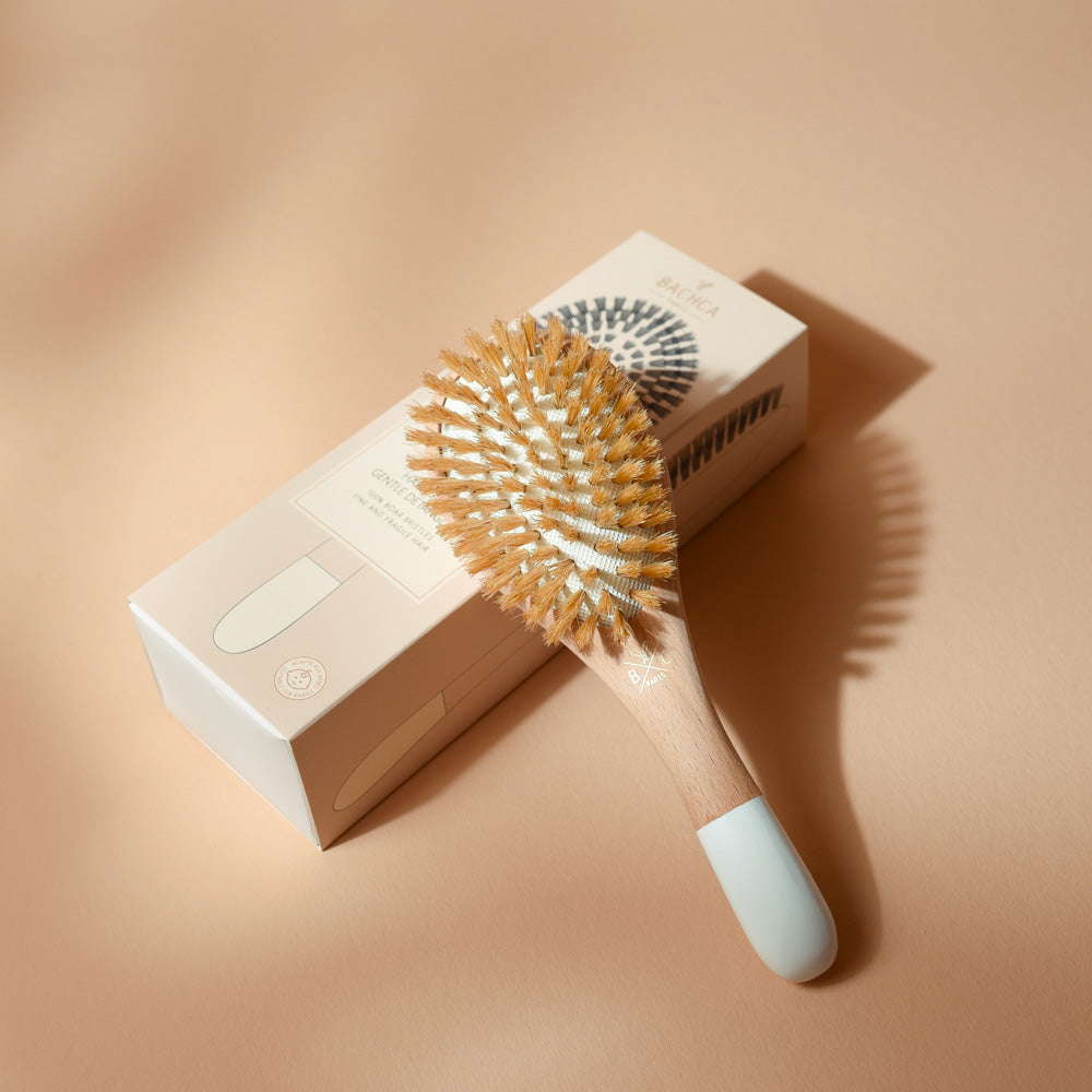 Brosse à cheveux Bachca grand format pour cheveux fins et fragiles, en bois de hêtre avec poils mixtes et manche blanc, posée sur sa boîte beige illustrée, sur fond sable.