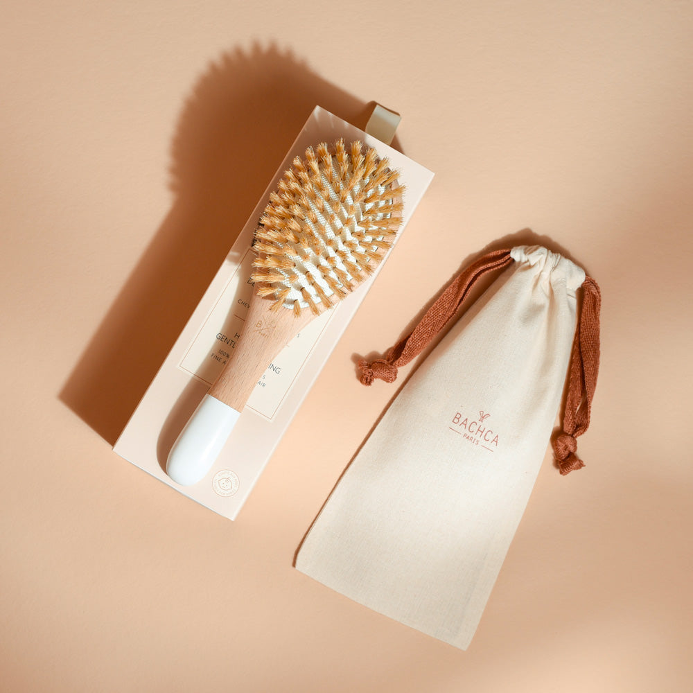 Brosse Bachca en bois clair et blanc, poils de sanglier blonds, posée sur son coffret beige avec pochon en coton à côté.