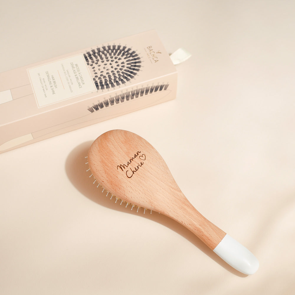 Une brosse à cheveux en bois clair Bachca avec embout blanc est posée sur un fond beige. Le dos de la brosse est gravé de l’inscription &
