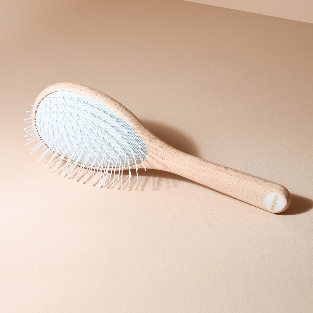 Brosse démêlante universelle Bachca à picots nylon blancs, posée en diagonale sur fond beige, avec manche en bois de hêtre brut et pastille nacrée au bout.