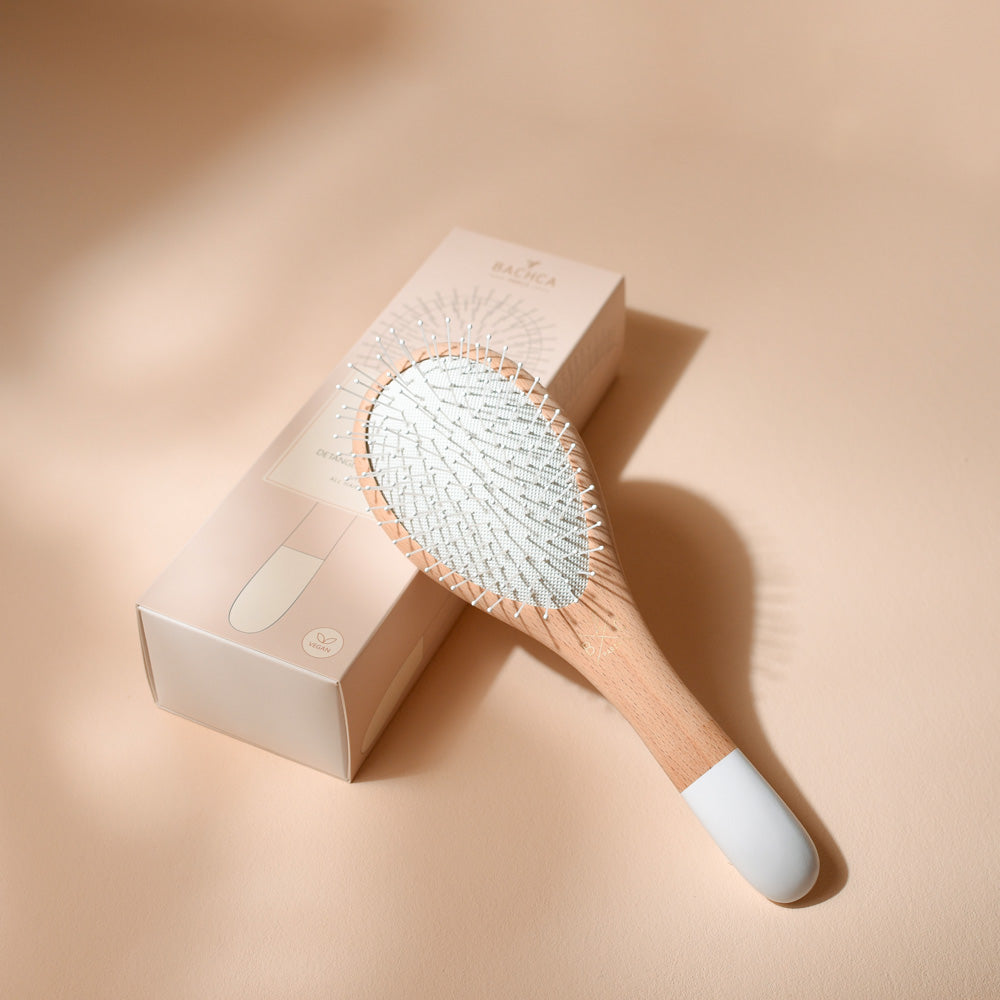Brosse Bachca démêlage et volume en bois de hêtre et manche blanc, posée sur son étui rose poudré.