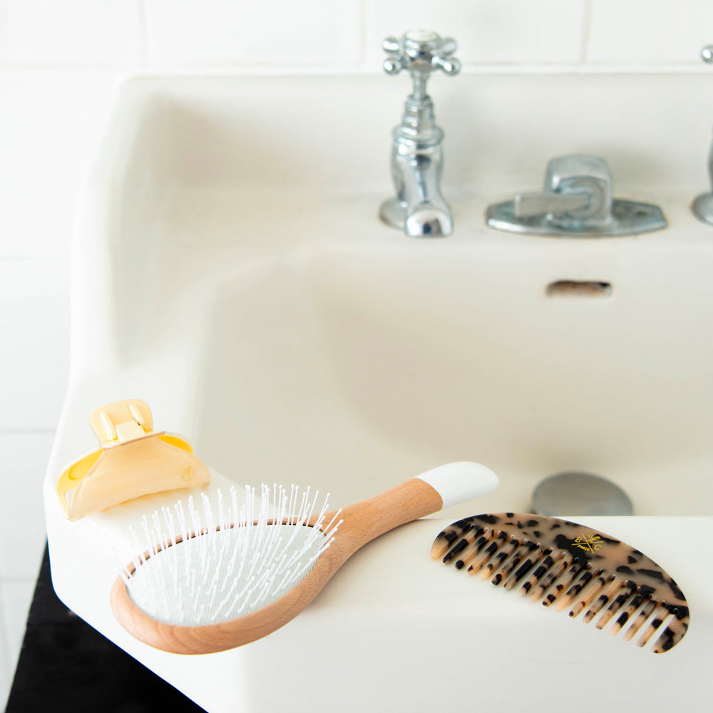 Accessoires capillaires Bachca posés sur un lavabo blanc : une brosse en bois à manche blanc, un peigne en acétate façon écaille et une pince à cheveux jaune pâle.