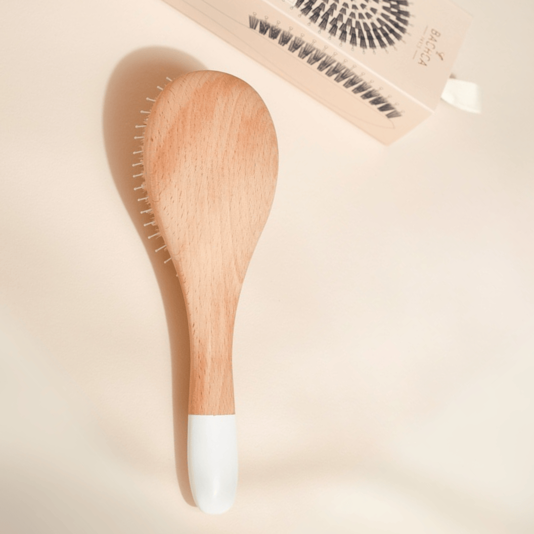 Brosse démêlante Bachca en bois de hêtre naturel avec embout blanc, posée à plat sur fond beige, accompagnée de son étui illustré.