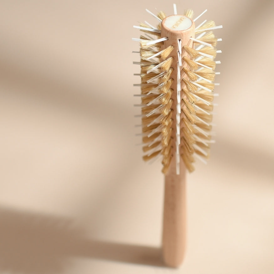 Brosse ronde de brushing Bachca en bois naturel posée verticalement, vue en perspective sur les rangées de poils mixtes en sanglier et en nylon, sur fond beige doux avec ombres marquées.