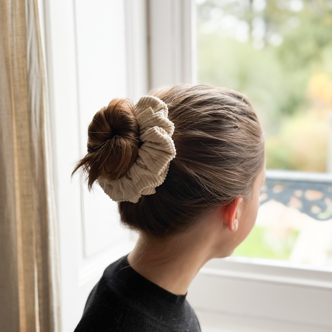 Chignon serré maintenu par un chouchou en velours côtelé beige Bachca, porté par une femme de dos regardant par la fenêtre.