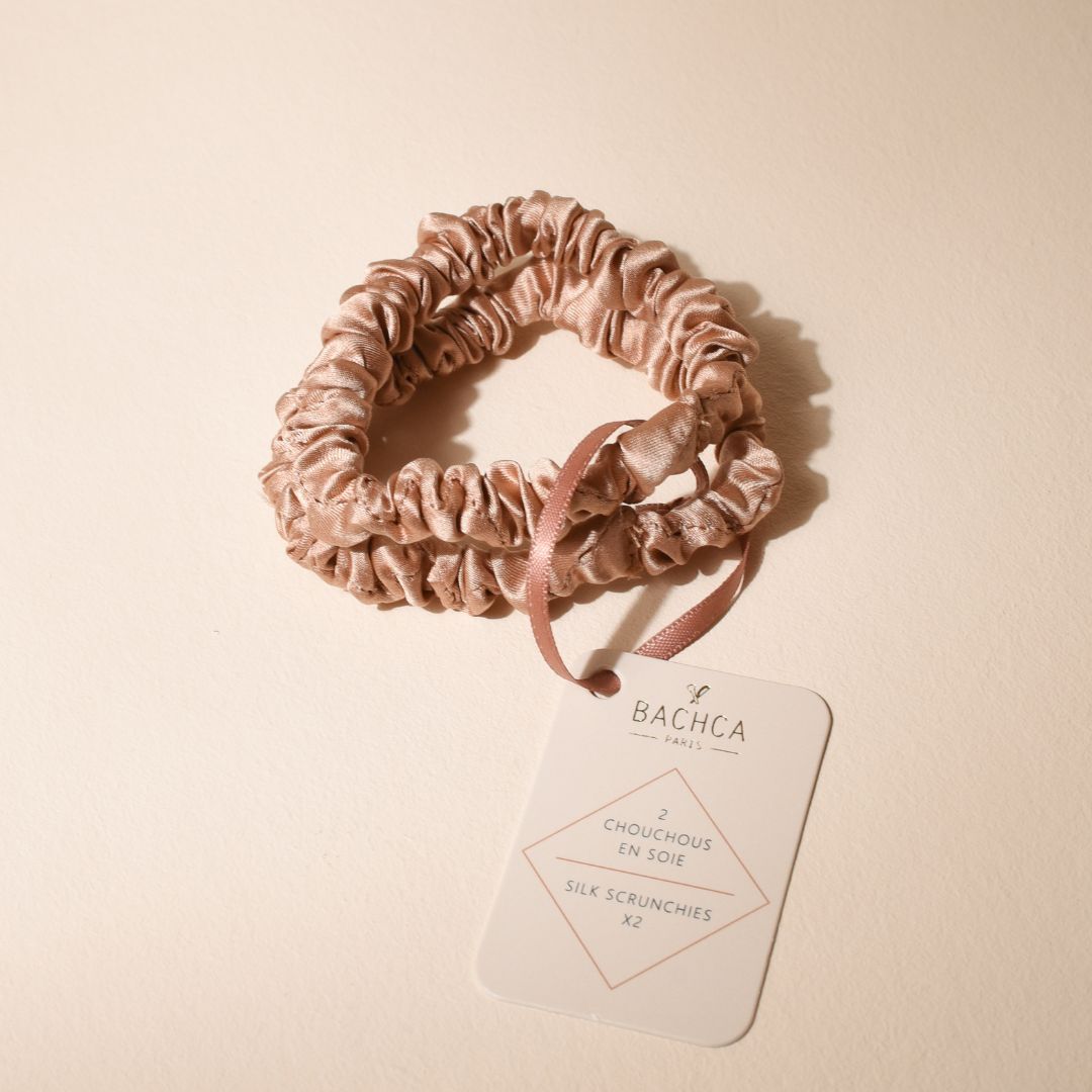 Deux chouchous en soie rose Bachca, maintenus ensemble par un ruban assorti et une étiquette blanche indiquant « 2 chouchous en soie – Silk Scrunchies x2 », posés sur un fond beige doux.