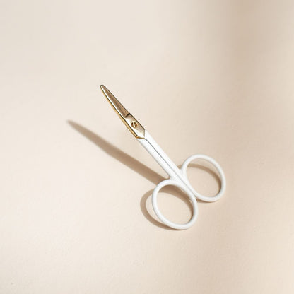 Ciseaux bébé Bachca au design ergonomique, manches blancs et lames dorées arrondies, posés sur un fond beige doux avec ombre portée.