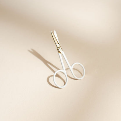 Ciseaux pour ongles bébé Bachca en métal doré et poignées blanches, posés debout sur un fond beige avec une ombre douce.