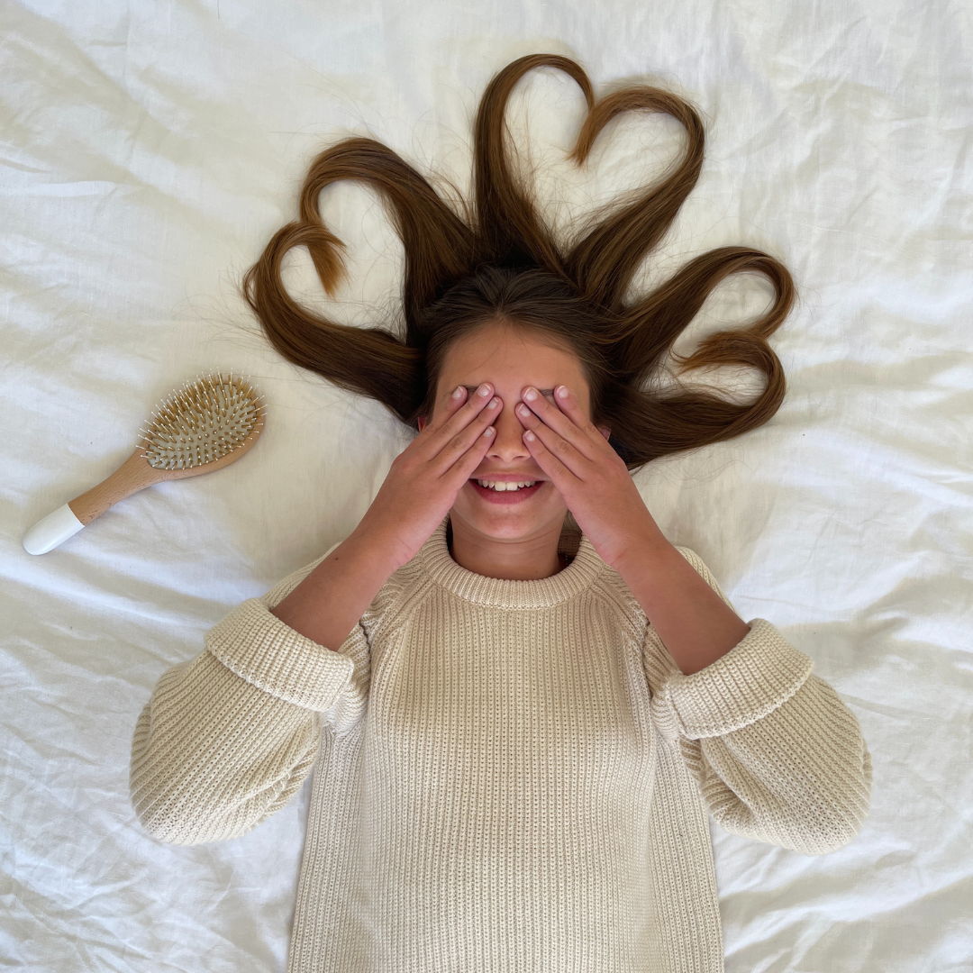 Petite fille allongée sur un lit, cheveux formant des cœurs, avec une brosse Bachca posée à côté, ambiance douce et ludique.