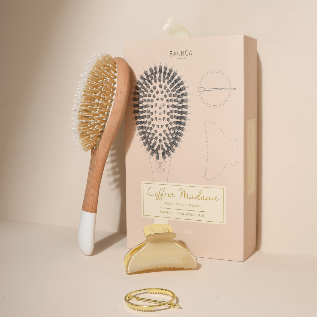 Coffret Madame Bachca debout, mis en scène avec une brosse en bois à picots sanglier et nylon, une pince crème et une barrette ronde dorée, sur fond beige.